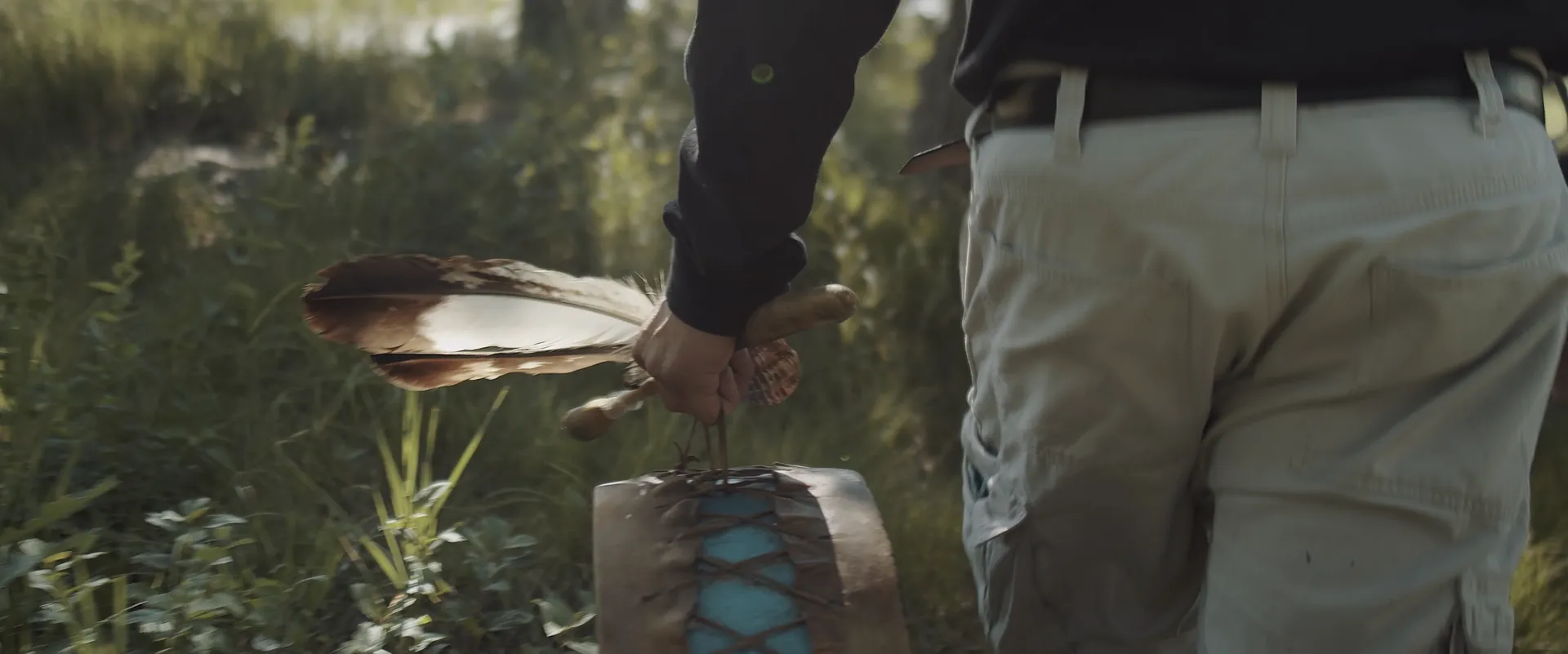 A hand carrying a feather and a drum through tall grass